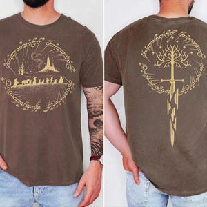 a man wearing a brown t-shirt with a graphic design featuring a castle, a tree, and a sword. The design is symmetrical, with the castle on the left and the sword on the right. The man has tattoos on his arms, and the back of the t-shirt displays the same design.
