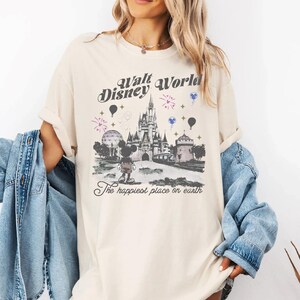 A woman wearing a white t-shirt with a graphic design of Disney World, including a castle, balloons, and the text "Disney World" and "The happiest place on earth".