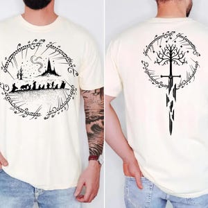 a man wearing a white t-shirt with a graphic design featuring a castle, a tree, and a sword. The design is circular and includes text and symbols.