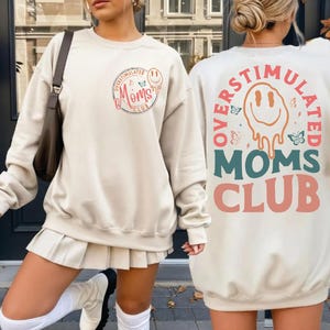 May include: Cream-colored sweatshirt with the text "OVERSTIMULATED MOMS CLUB" in a circular design on the front and back. The front design includes a smiling face and butterflies. The back design features a melting smiley face.