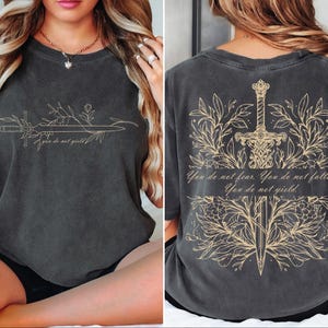 May include: Dark gray t-shirt with a gold sword and floral design. The front features a sword with the text "you do not yield". The back displays a sword surrounded by leaves and the text "You do not fear. You do not falter. You do not yield."
