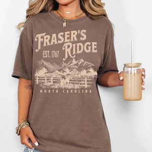A woman wearing a brown t-shirt with the text "Fraser's Ridge" and "Est. 1767" printed on it, along with a mountain landscape. She is holding a glass of what appears to be a beverage, and there is a necklace visible around her neck.