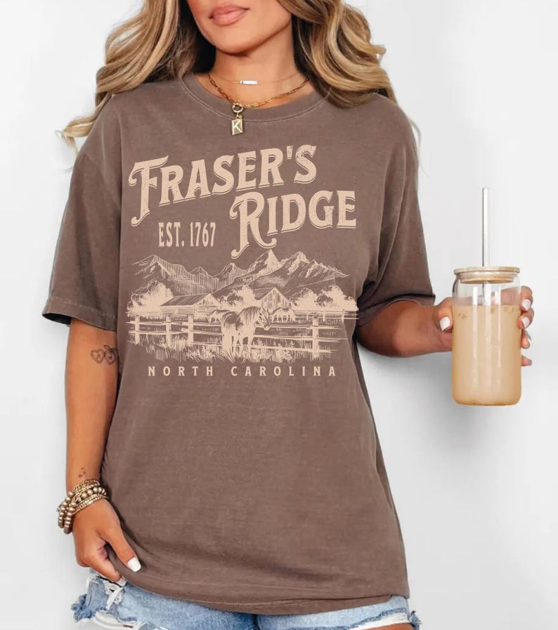 A woman wearing a brown t-shirt with the text "Fraser's Ridge" and "Est. 1767" printed on it, along with a mountain landscape. She is holding a glass of what appears to be a beverage, and there is a necklace visible around her neck.
