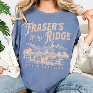 A woman wearing a blue t-shirt with the text "Fraser's Ridge" and "Est. 1767 North Carolina" printed on it.