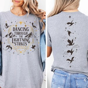 A woman wearing a gray t-shirt with a graphic design featuring a mountain range and the text "Dancing through the Lightning Strikes".