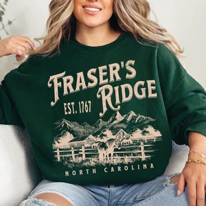 A woman wearing a green sweatshirt with a graphic design of a mountain range and the text "Fraser's Ridge" printed on it.