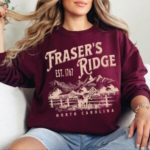 A woman wearing a burgundy sweatshirt with a graphic design of a mountain range and the text "Fraser's Ridge" printed on it.