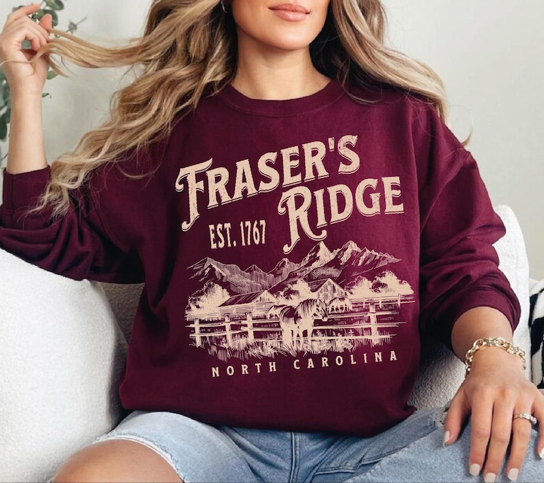 A woman wearing a burgundy sweatshirt with a graphic design of a mountain range and the text "Fraser's Ridge" printed on it.
