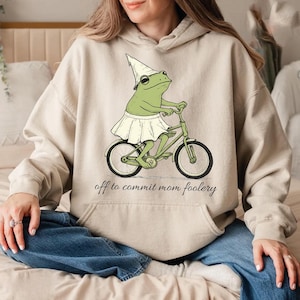 May include: Beige hoodie with a whimsical graphic of a green frog riding a bicycle, wearing a skirt and hat. The text "off to commit mom foolery" is printed below the graphic. The hoodie has long sleeves and a front pocket.