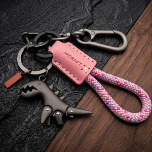 Könnte beinhalten: Ein Schlüsselanhänger mit einem grauen Dinosaurier-Flaschenöffner, einem rosa Lederanhänger mit dem Text "H-CRAFT" und einer rosa, blauen und weißen Seilschlaufe. Er enthält Metallkarabiner und Schlüsselringe. Der Hintergrund ist eine Mischung aus Dunkelgrau und braunem Holz.