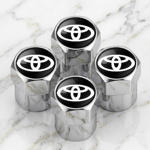 Op de afbeelding: Vier verchroomde ventieldopjes met een zwart midden en een wit Toyota-logo. De doppen zijn zeshoekig en in een cluster gerangschikt, waardoor ze licht reflecteren. De achtergrond is een marmerachtig oppervlak.