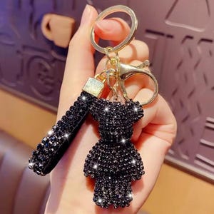 Op de afbeelding: Een sleutelhanger met een beervormige bedel en een bijpassende rechthoekige riem, beide bedekt met kleine, sprankelende zwarte kristallen. Het beslag is goudkleurig en bestaat uit een ronde splitring en een karabijnhaak voor bevestiging.