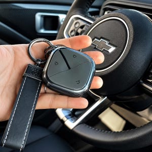 Peut inclure: Un étui de clé de voiture gris foncé de forme carrée avec des coins arrondis et trois icônes de fonction, attaché à une lanière en cuir noir avec des coutures blanches et un anneau métallique. La lanière porte le texte Good Luck sur le fermoir.