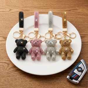 Puede incluir: Cuatro llaveros con forma de oso con superficies incrustadas de pedrería en negro, rosa, plata y oro. Cada llavero incluye una correa a juego cubierta de pedrería, un mosquetón de metal dorado y una anilla circular para llaves.