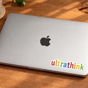 May include: A silver laptop with a black Apple logo and a colourful "ultrathink" sticker on a wooden desk. A cup of coffee, a small plant, glasses, a notepad, and a pen are also on the desk.