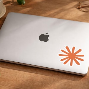 May include: A closed silver laptop with a black Apple logo. An orange starburst sticker with a white border is on the laptop. The laptop is on a wooden surface with a cup, glasses, a notepad, and a pen.