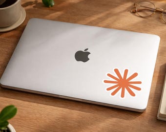 May include: A closed silver laptop with a black Apple logo. An orange starburst sticker with a white border is on the laptop. The laptop is on a wooden surface with a cup, glasses, a notepad, and a pen.