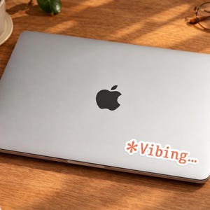 May include: A silver laptop with a black Apple logo on the lid. A sticker with the text "*Vibing..." in red is on the lower right corner. The laptop is on a wooden desk, with a cup of coffee, glasses, a notepad, and a pen.