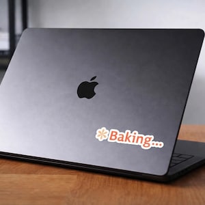 May include: A black laptop with the Apple logo on the lid. A sticker with the word "Baking..." in red and white is on the laptop. The laptop is open on a wooden surface.