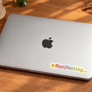 May include: A silver laptop with a black Apple logo and a colourful sticker that says "Manifesting...". The laptop is on a wooden surface, next to a cup of coffee, glasses, a notepad, and a pen.