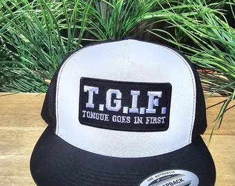 May include: Black and white trucker cap with a flat peak. The front panel is white with a black rectangle patch that reads "T.G.I.F. TONGUE GOES IN FIRST" in blue and white lettering. The cap has a black mesh back and a snapback closure.