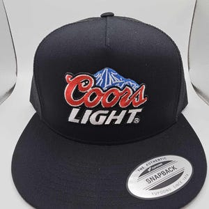 Puede incluir: Gorra snapback negra con visera plana. La gorra presenta el logotipo de Coors Light en bordado rojo, blanco y azul. Tiene una parte trasera de malla y un cierre a presión. La etiqueta en la visera dice "The Authentic Snapback Yupoong Since 1974."