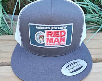 May include: A grey and white trucker cap with a flat peak. The front displays a rectangular patch with the text "America's Best Chew" and "Red Man Chewing Tobacco" in red and white. The cap has a snapback closure.