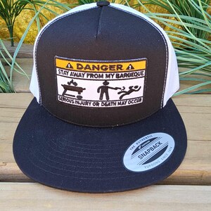 Op de afbeelding: Zwart-witte snapback pet met een platte klep. Het voorpaneel heeft een patch met de tekst "DANGER STAY AWAY FROM MY BARBEQUE SERIOUS INJURY OR DEATH MAY OCCUR". De pet heeft een witte mesh achterkant en een zwarte klep.