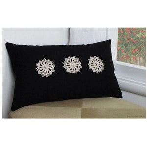 May include: Black rectangular decorative pillow with three cream-colored, circular, crocheted floral designs. The pillow is placed on a beige surface near a window with a view of red flowers.