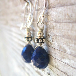 May include: A pair of silver earrings with blue gemstone drops. The earrings feature a silver bead with a textured surface and a silver hook.
