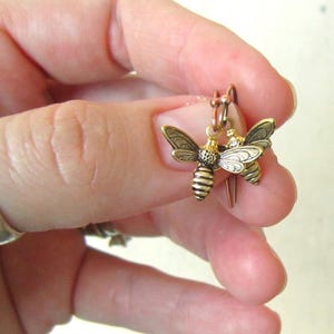 May include: A pair of small, gold-toned bee-shaped earrings featuring detailed wings and a striped abdomen. The earrings are suspended from copper-colored metal ear wires, showcasing a metallic finish with intricate textures on the insect bodies.