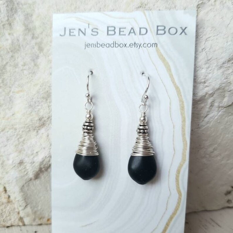 May include: Black gemstone earrings with silver wire wrapped around the stone and silver beads at the top of the wire. The earrings are hanging from silver hooks.