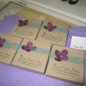 K&ouml;nnte beinhalten: F&uuml;nf kleine braune Pappkartons mit rustikalem Jute- und t&uuml;rkisfarbenem Banddesign. Jede Schachtel hat eine lila Blume mit einem Perlenmittelpunkt und den Text "Jen's Bead Box Rustikale inspirierte handgefertigte Schmuckst&uuml;cke www.jensbeadbox.etsy.com" auf der Vorderseite.