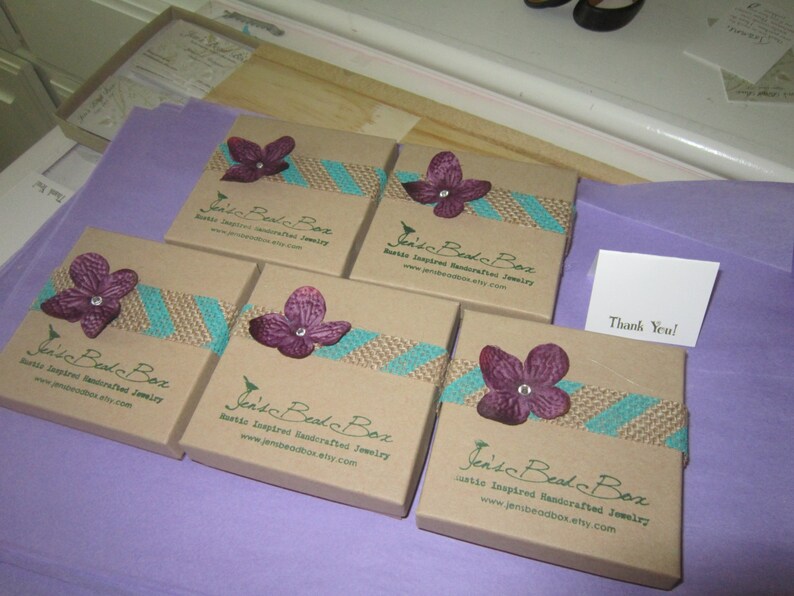 K&ouml;nnte beinhalten: F&uuml;nf kleine braune Pappkartons mit rustikalem Jute- und t&uuml;rkisfarbenem Banddesign. Jede Schachtel hat eine lila Blume mit einem Perlenmittelpunkt und den Text "Jen's Bead Box Rustikale inspirierte handgefertigte Schmuckst&uuml;cke www.jensbeadbox.etsy.com" auf der Vorderseite.