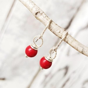 May include: A pair of red bead earrings with silver-colored hooks and accents. The round red beads are suspended from the hooks, creating a simple yet elegant design. The earrings are displayed on a small, light-colored branch.