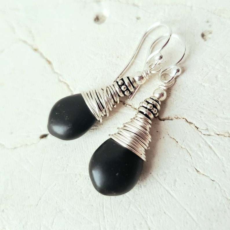 May include: A pair of dangle earrings with black gemstone drops wrapped in silver wire. The earrings have silver hooks.