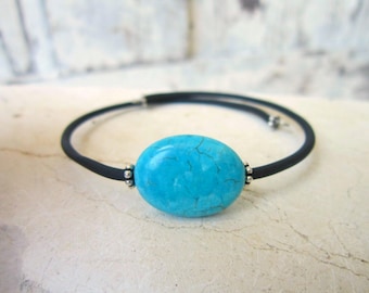 May include: A bracelet featuring a large, oval-shaped turquoise stone with dark veining, centered on a thin, flexible black band. The stone is flanked by small, round silver-toned metal beads that connect the turquoise to the black cord.