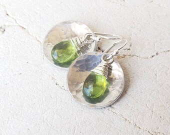 Peut inclure: Une paire de boucles d'oreilles en argent avec des gouttes de pierres précieuses vertes. Les boucles d'oreilles présentent des disques en argent martelé avec une pierre précieuse verte enveloppée de fil qui pend de chaque côté.