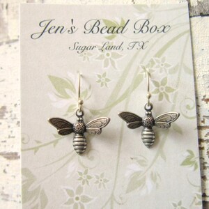 May include: A pair of silver-toned bee-shaped dangle earrings with hook backings, displayed on a light beige card featuring the text Jen's Bead Box Sugar Land, TX and a subtle white floral pattern printed on the card surface.