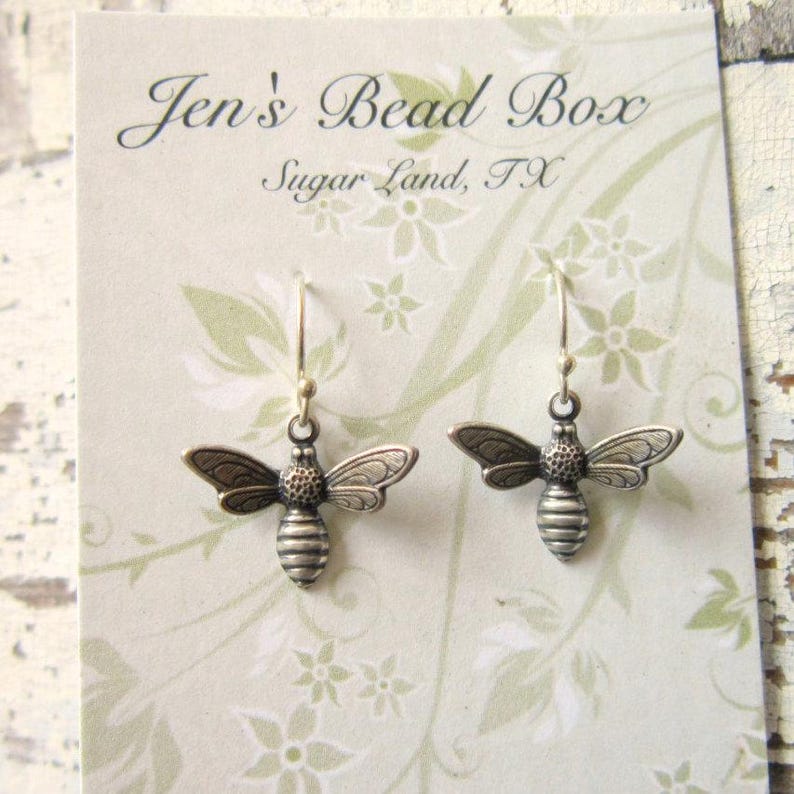 May include: A pair of silver-toned bee-shaped dangle earrings with hook backings, displayed on a light beige card featuring the text Jen's Bead Box Sugar Land, TX and a subtle white floral pattern printed on the card surface.