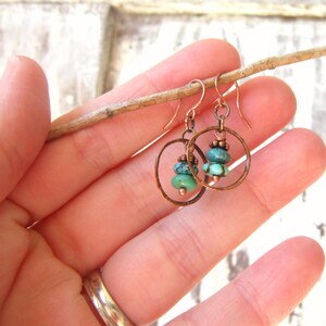 May include: A pair of copper hoop earrings with turquoise gemstone beads. The earrings are hanging from a twig held in a hand.
