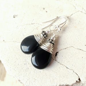 May include: A pair of black teardrop-shaped earrings. The earrings feature a silver wire wrap design around the top of the black stone and a silver hook. The earrings are displayed on a white surface.