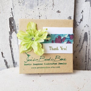 May include: A small brown box with a green flower and a blue and purple ribbon. The box has the text "Jen's Bead Box" and "Rustic Inspired Handcrafted Jewelry" printed on it. The box also has a white card with the text "Thank You!" printed on it.
