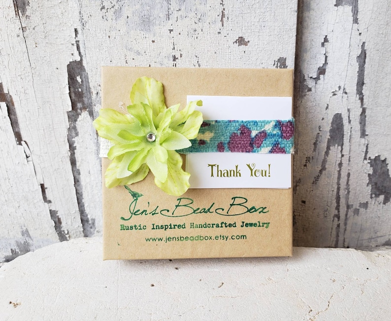 May include: A small brown box with a green flower and a blue and purple ribbon. The box has the text "Jen's Bead Box" and "Rustic Inspired Handcrafted Jewelry" printed on it. The box also has a white card with the text "Thank You!" printed on it.