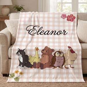 May include: A soft throw blanket featuring a light pink and white gingham pattern. The name Eleanor is printed in black script. Below the name are illustrations of a black and white cat, a yellow duck, a brown bear, an owl, and a chicken.
