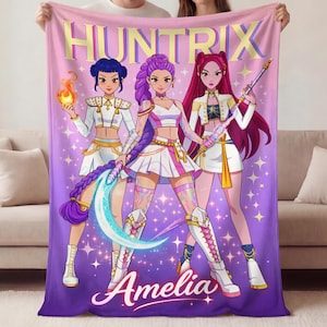 May include: A plush blanket featuring a vibrant illustration of three anime-style characters. The blanket has a gradient background of pink and purple with the word "HUNTRIX" in gold lettering. The name "Amelia" is written in cursive at the bottom.