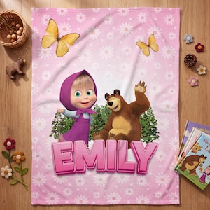 May include: A soft pink fleece blanket featuring a repeating white daisy pattern, two yellow butterflies, and a graphic of Masha and the Bear standing in front of green pine trees. The name EMILY is printed in large, bold, pink 3D-style letters.