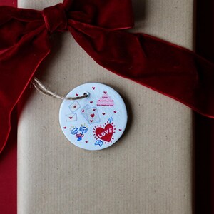 Puede incluir: Un regalo envuelto en papel marrón, atado con un gran lazo de terciopelo rojo oscuro. Una etiqueta redonda blanca con un corazón que dice "LOVE" y otras ilustraciones está sujeta con hilo.