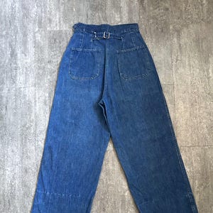 May include: Blue denim wide-leg pants with a high waist and belt loops. The back features two square pockets and a belt with a silver buckle. The hem of the pants has a white accent line.