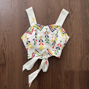 May include: A white sleeveless crop top featuring a colorful geometric pattern in red, yellow, blue, and green. The garment has wide shoulder straps, a sweetheart neckline, and a tie-front closure at the hem, showcasing a repeating abstract motif.
